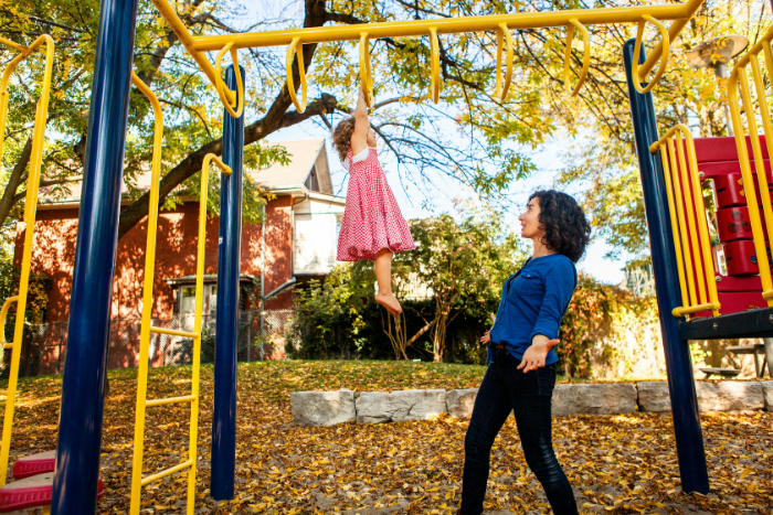 Child in a polka dot dress hanging from playground monkey bars while a parent watches, autumn leaves on the ground