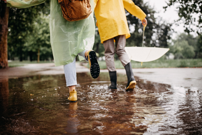 Parent and child in raincoats and rain boots splashing through a puddle on a wet park path